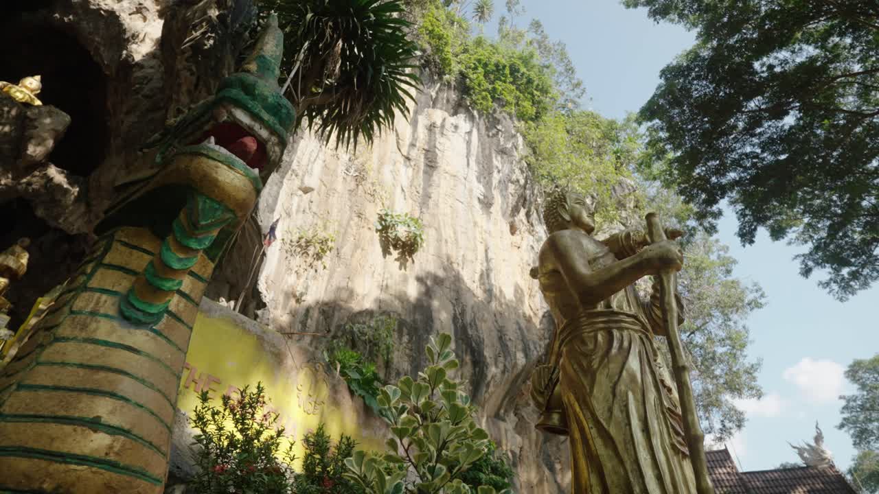 Golden Buddha and serpent statues stand beneath steep cliffs in Wat Tham Phra