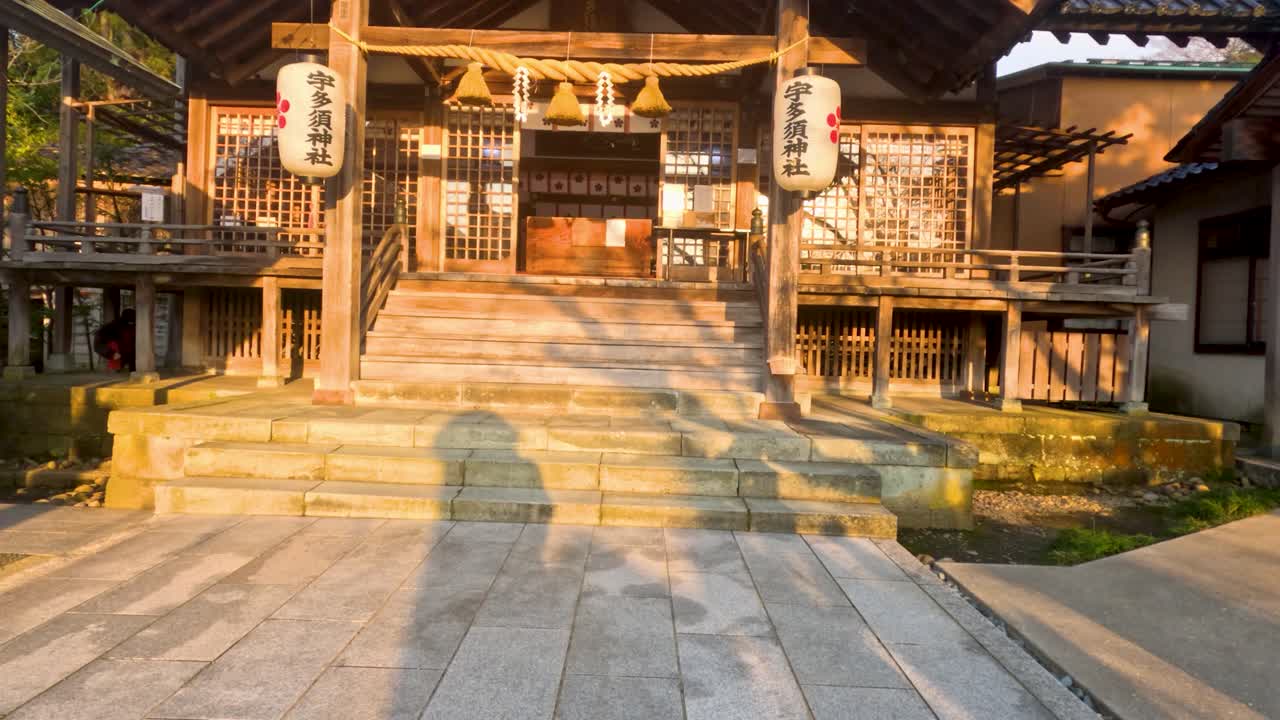 温かい夕焼けの光が、石畳に長い影を落としながら伝統的な木造の神社を照らしています。