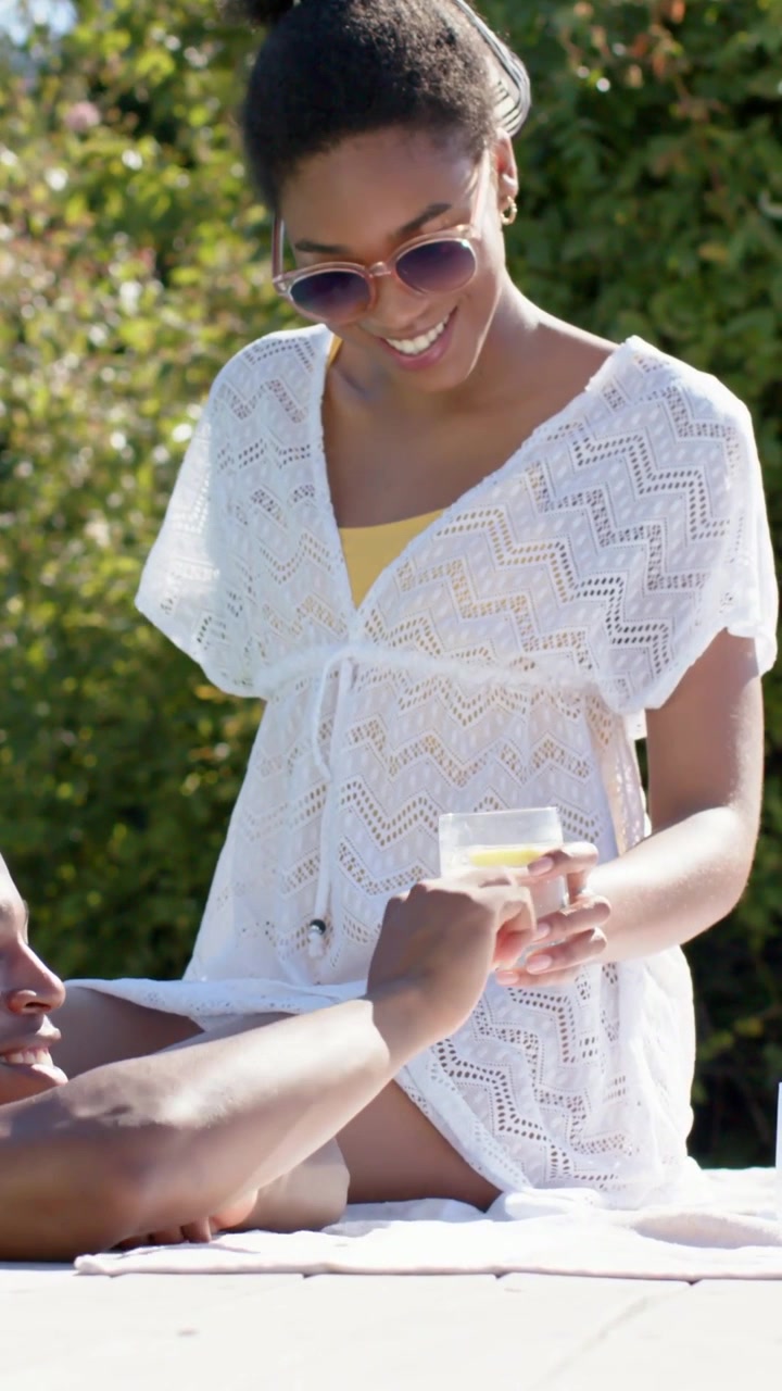 Vertical video: Smiling woman in swimsuit leaning forward handing lemon glass pool for him to drink