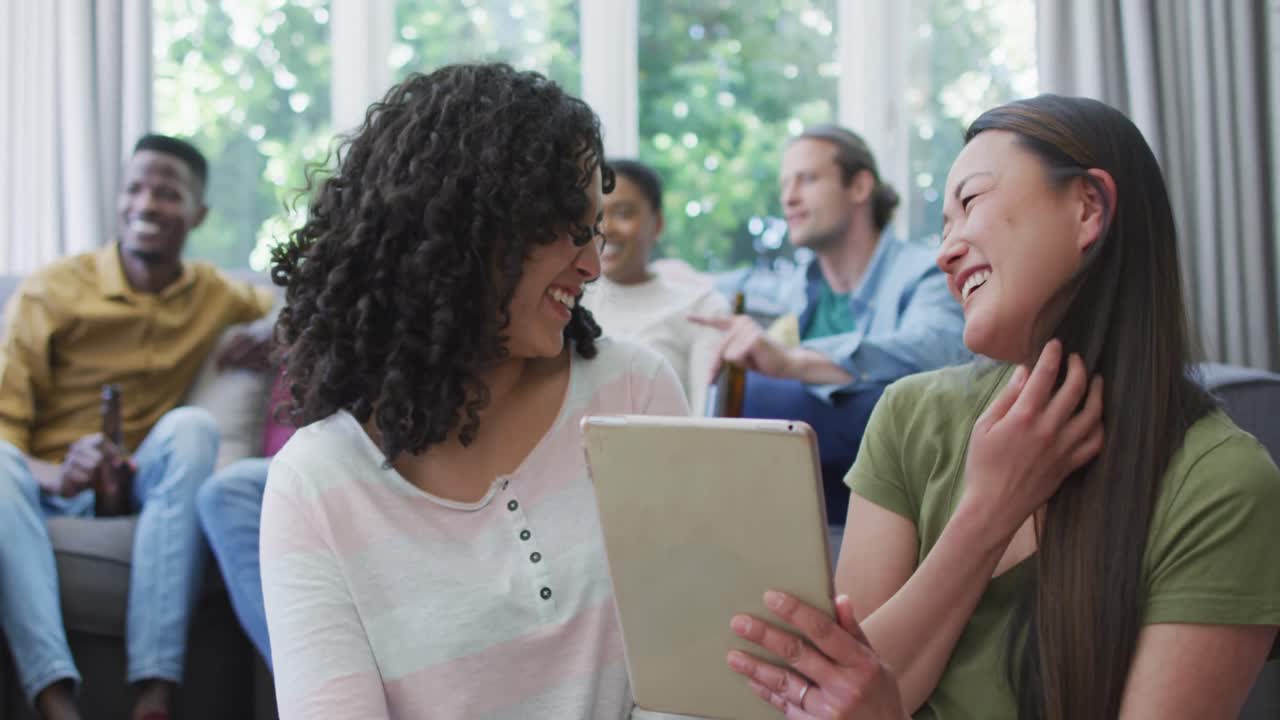 amigas diversas mirando la tableta y riendo, con amigos en la sala de estar