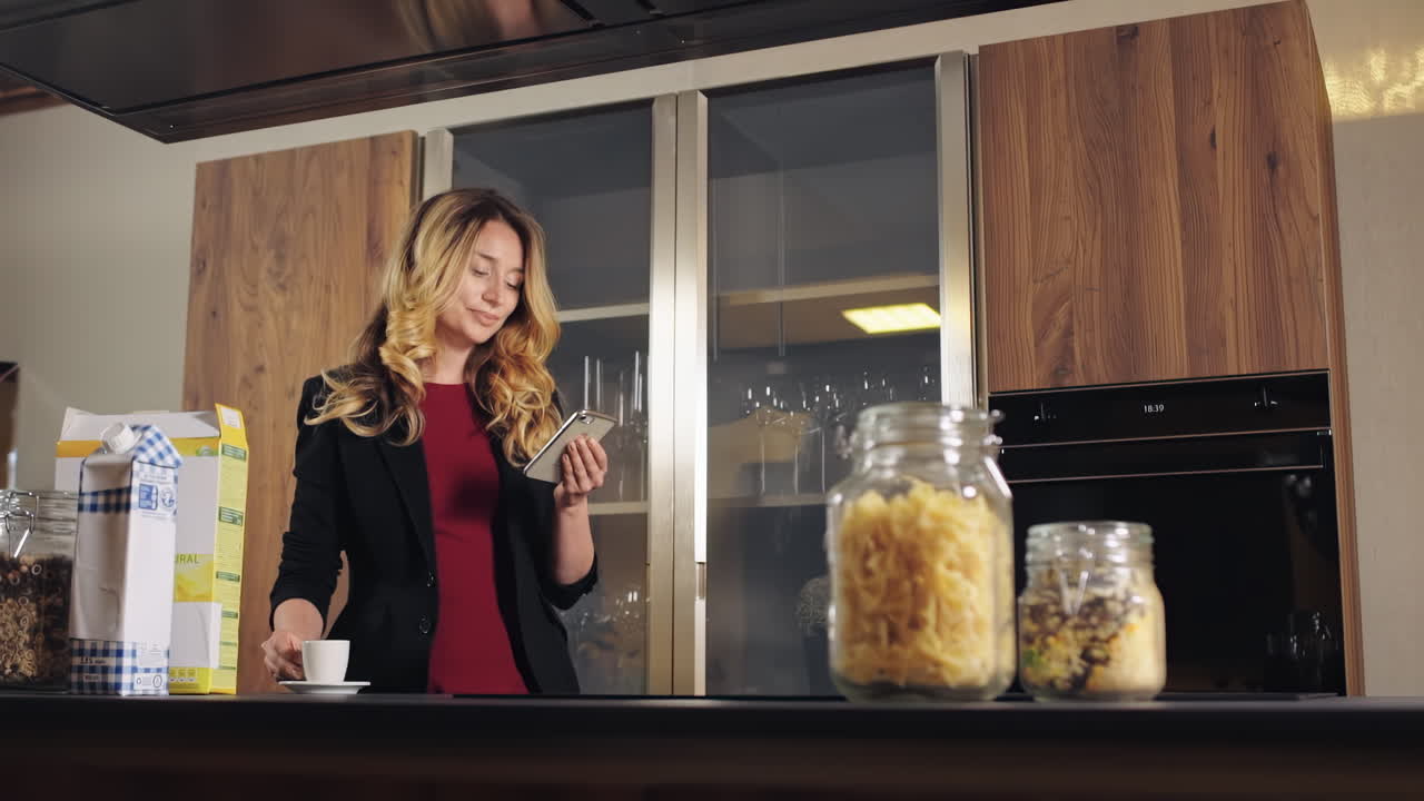 CHISINAU, MOLDOVA - SEPTEMBER 25, 2018: A laughing caucasian woman with blonde hair in jacketlooking at the phone on a kitchen. Table with food and cup of coffee