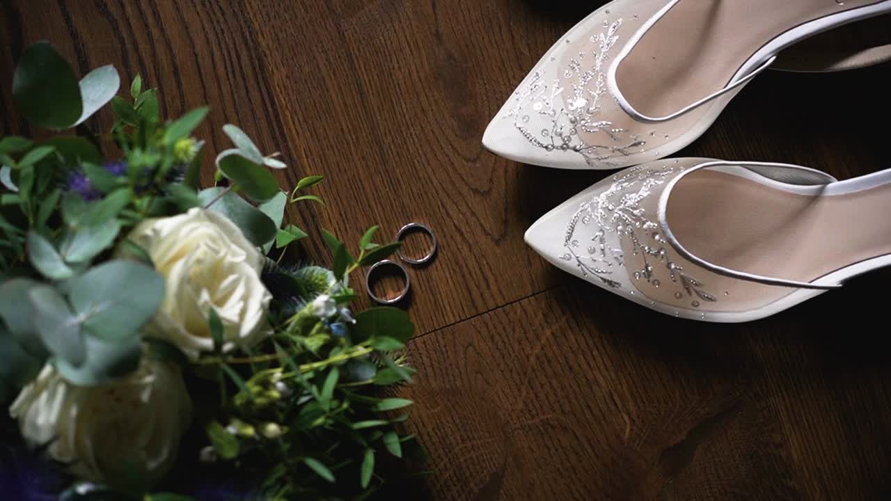 Top view of the wedding rings, bridal shoes, bridal long dress and flower bouquet on a dark wooden surface, rotating shot
