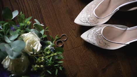 top view of the wedding rings, bridal shoes, bridal long dress and flower bouquet on a dark wooden surface, rotating shot