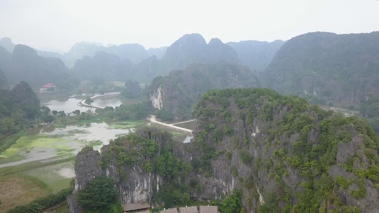 Cinematic drone footage of Tam Coc surrounded by majestic limestone peaks in misty and foggy weather. Ricefields in the wild jungle in the spring with boat tour canal in, Ninh Binh, Vietnam