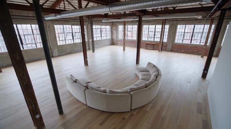 La telecamera inizia una panoramica, inquadrando un divano semicircolare in un loft industriale, rivelando le travi a vista