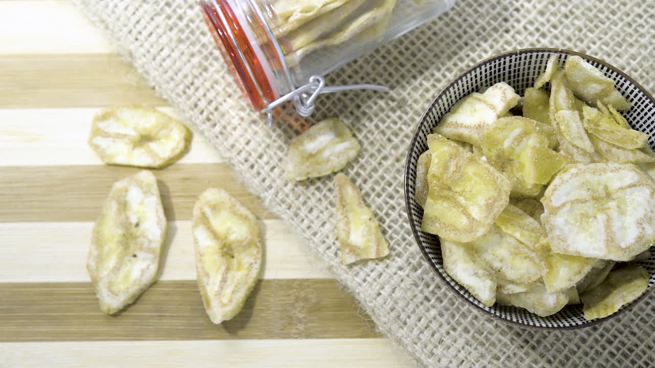 toma panorámica lenta vista de arriba hacia abajo sobre un tazón de cerámica de un frasco de vidrio lleno de una mezcla de castañas con pasas sobre una mesa a rayas con tela de arpillera