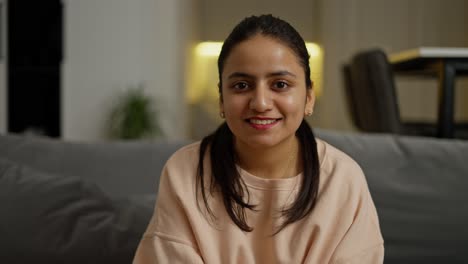 Portrait-of-a-happy-brunette-girl-with-brown-skin-in-a-beige-jacket-who-sits-on-a-gray-sofa-in-a-modern-apartment-during-the-day.-Portrait-of-a-happy-brunette-girl-in-a-modern-apartment-during-the-day