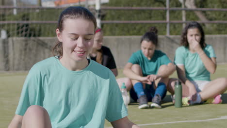 happy teenaged girl holding football in front of camera, while her teammates relax behind 1