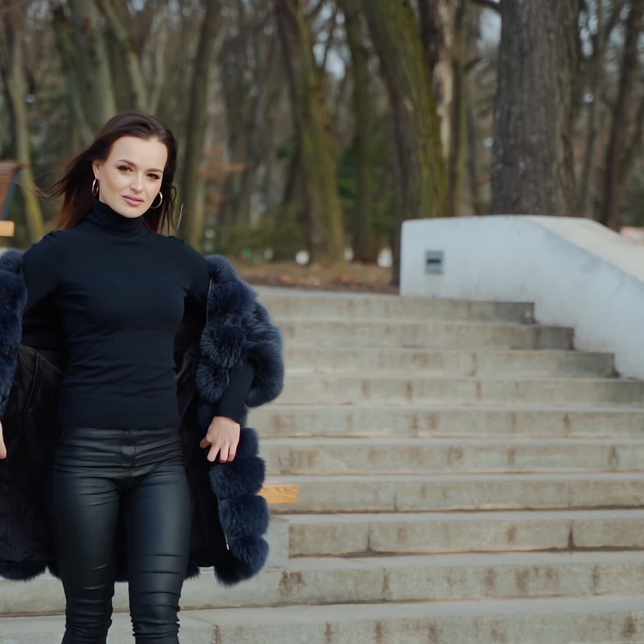 Beautiful model posing on camera in autumn park. Young woman in black fur coat walking and looking at camera. Slow motion.