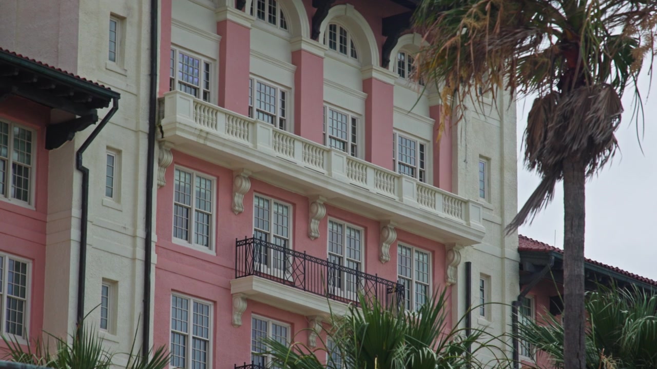 Foto em grande angular de uma fachada de hotel rosa pastel no Texas, emoldurada por palmeiras balançando. A luz suave do dia realça detalhes arquitetônicos e vegetação tropical para um ambiente de resort de luxo.