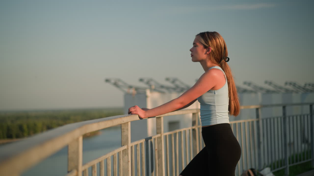 joven dama descansando la mano en la barandilla de hierro mientras mira pensativamente en la distancia bajo la cálida luz del sol, con la pierna derecha avanzada, el fondo presenta vegetación distante y estructuras industriales