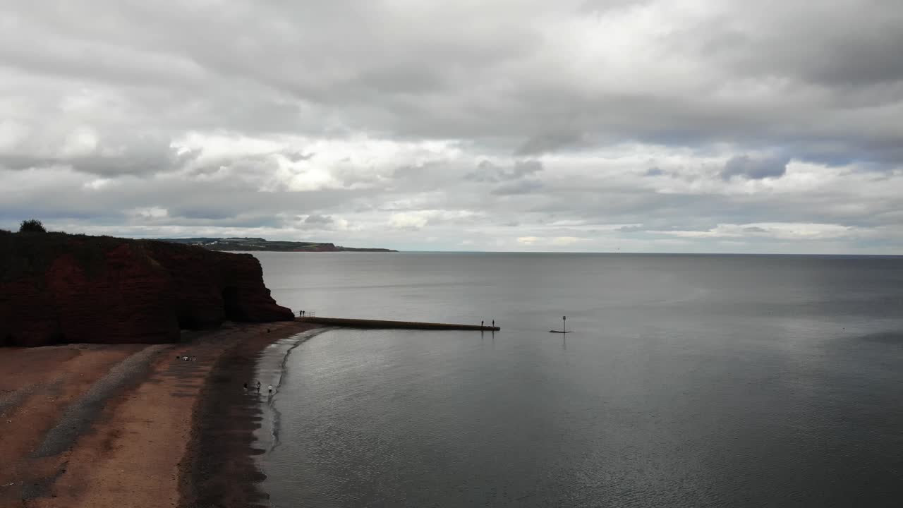 Aerial view of Red cliffs overlook calm waters and a peaceful beach under cloudy skies. rising shot