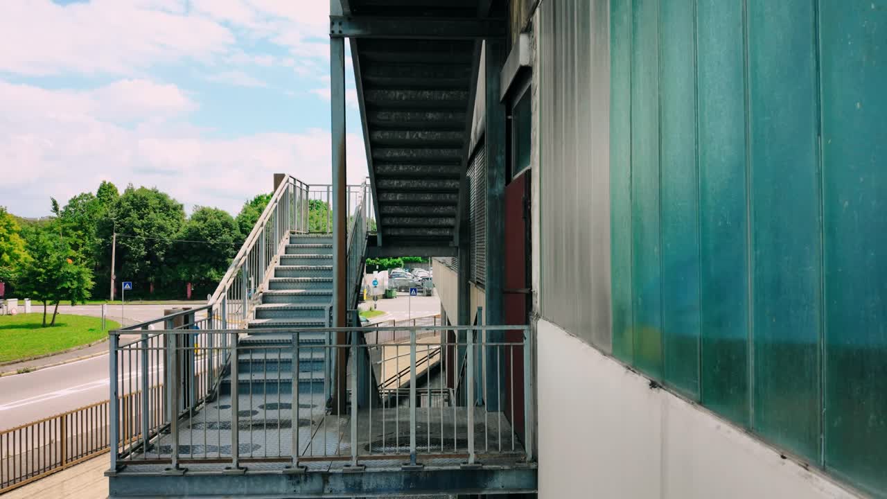 Vista dal basso di una scala metallica industriale sul lato di un edificio con pannelli di vetro blu nella città di Bergamo. Architettura urbana caratterizzata da ringhiere in acciaio e scale in cemento vicino al traffico stradale intenso.