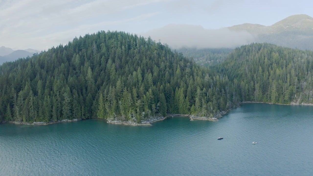 Un promontorio insulare coperto di alberi si erge sopra un'insenatura blu mentre un drone sorvola, rivelando due piccole imbarcazioni e montagne sfocate in lontananza