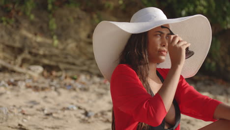 Modelo-De-Chica-Latina-Se-Sienta-Casualmente-Para-Ponerse-Gafas-De-Sol-Mientras-Se-Broncea-En-La-Playa-Bajo-El-Sol-Brillante,-Inclinación