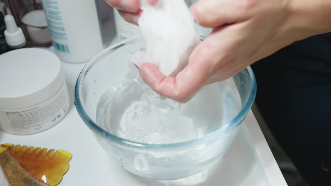 Hand view of facialist dipping cotton pad into glass bowl of purified water on white trolley beside skincare bottles, then gently pressing water from pad over facial bed in clinic room