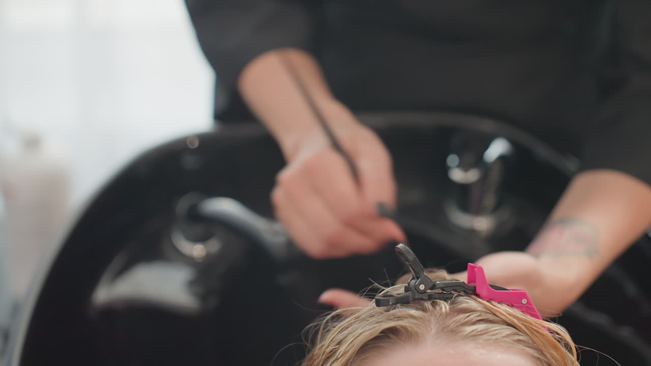 Lavaggio dei capelli in un salone con lavabo, parrucchiere tatuato deterge delicatamente il cuoio capelluto della cliente sopra il lavabo nero, clip rosa che fissa i capelli, schiuma cremosa e risciacquo caldo, cliente distesa che si gode la cura professionale e la tranquillità
