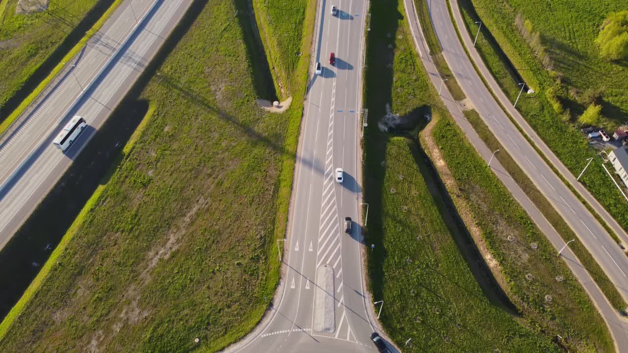 Drone top down shows Katlakalns highway interchange with lanes, ramps, roundabout, road markings, single car, springtime fields, shadows of lampposts, geometric infrastructure pattern