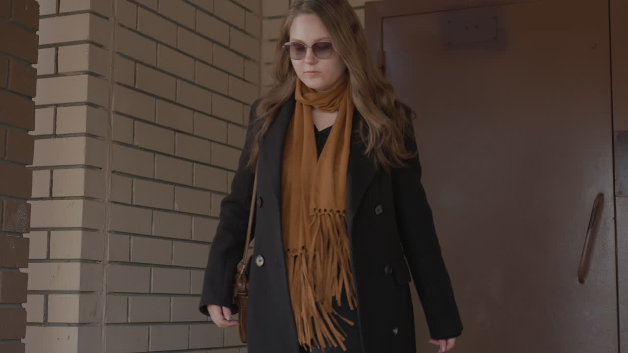 갈색 스카프와 갈색 가방을 메고 금발 머리를 휘날리며 선글라스를 낀 세련된 젊은 여성이 벽돌 출입구를 지나가는 모습이에요. 비즈니스 분위기와 차분한 자신감이 느껴지고, 은은한 햇빛 색감과 분위기를 담아낸 클로즈업 미드 샷이에요.