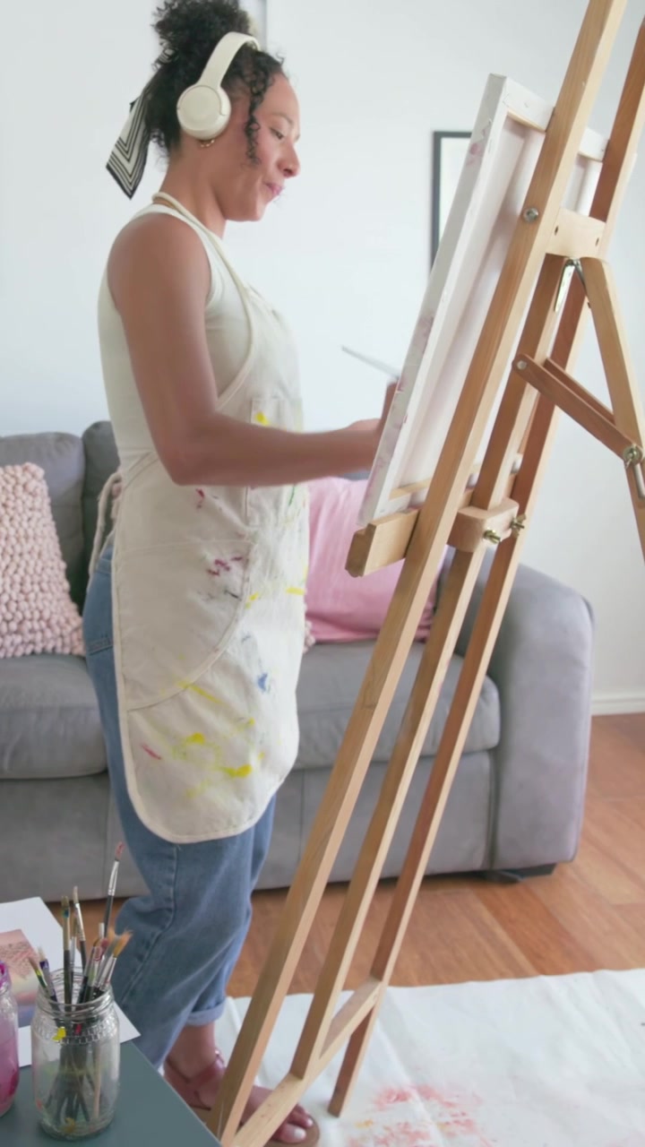 Video verticale: Donna con grembiule da pittore e telefoni, che sceglie i colori dalla tavolozza e aggiunge dettagli su un cavalletto a casa.