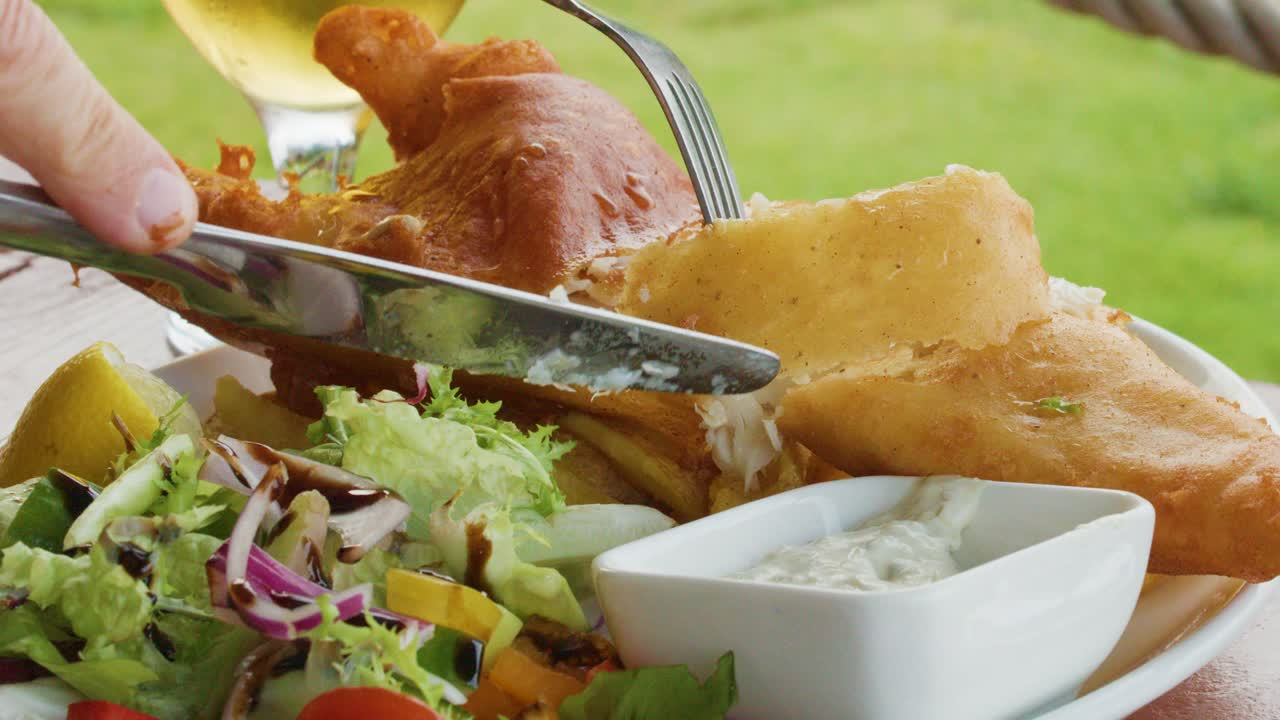 A person uses a fork and knife to cut into crispy battered fish, served with salad, tartar sauce, and beer on a sunny outdoor table