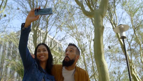 Vista-De-ángulo-Bajo-De-Una-Joven-Pareja-De-Raza-Mixta-Haciendo-Clic-En-Selfie-Con-Teléfono-Móvil-En-La-Ciudad-4k