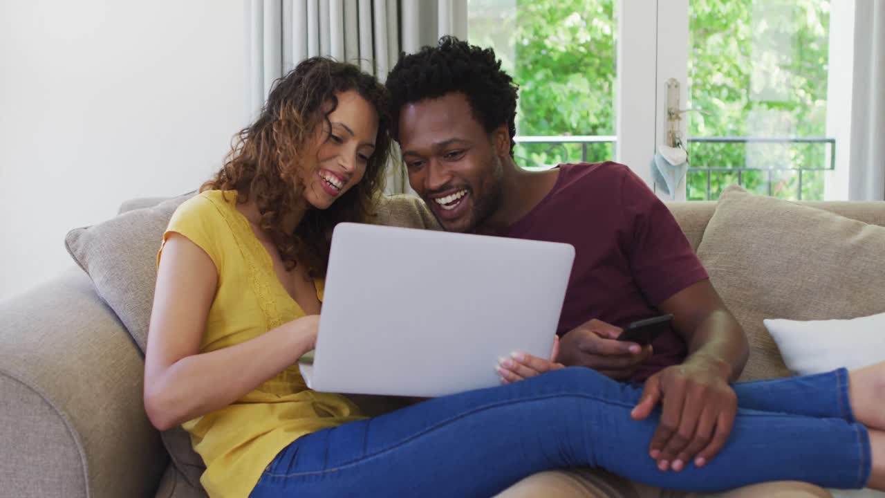 feliz pareja biracial sentada en el sofá con una computadora portátil y riendo