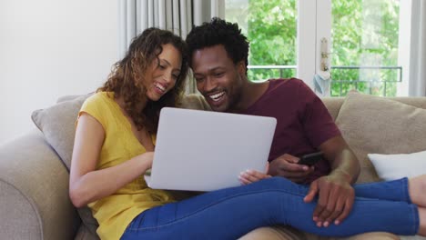 Happy-biracial-couple-sitting-on-sofa-with-laptop-and-laughing