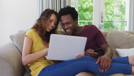 Feliz-Pareja-Birracial-Sentada-En-El-Sofá-Con-Una-Computadora-Portátil-Y-Riendo