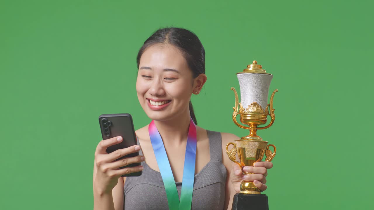 Close Up Of Asian Woman With A Gold Medal Holding A Gold Trophy And Using Smartphone On Green Screen Background In The Studio