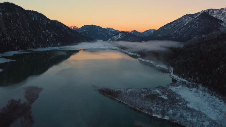 Flusstal der bayerisch-österreichischen Alpen mit frischem Wasser am Sylvensteinspeicher bei Sonnenuntergang, winterlich verschneitem Flussbett entlang von Bäumen, Wald und Bergen.