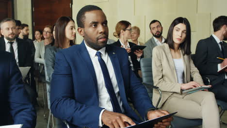 African-American-young-businessman-in-formal-clothes-sitting-at-the-business-conference,-typing-on-the-tablet-and-listening