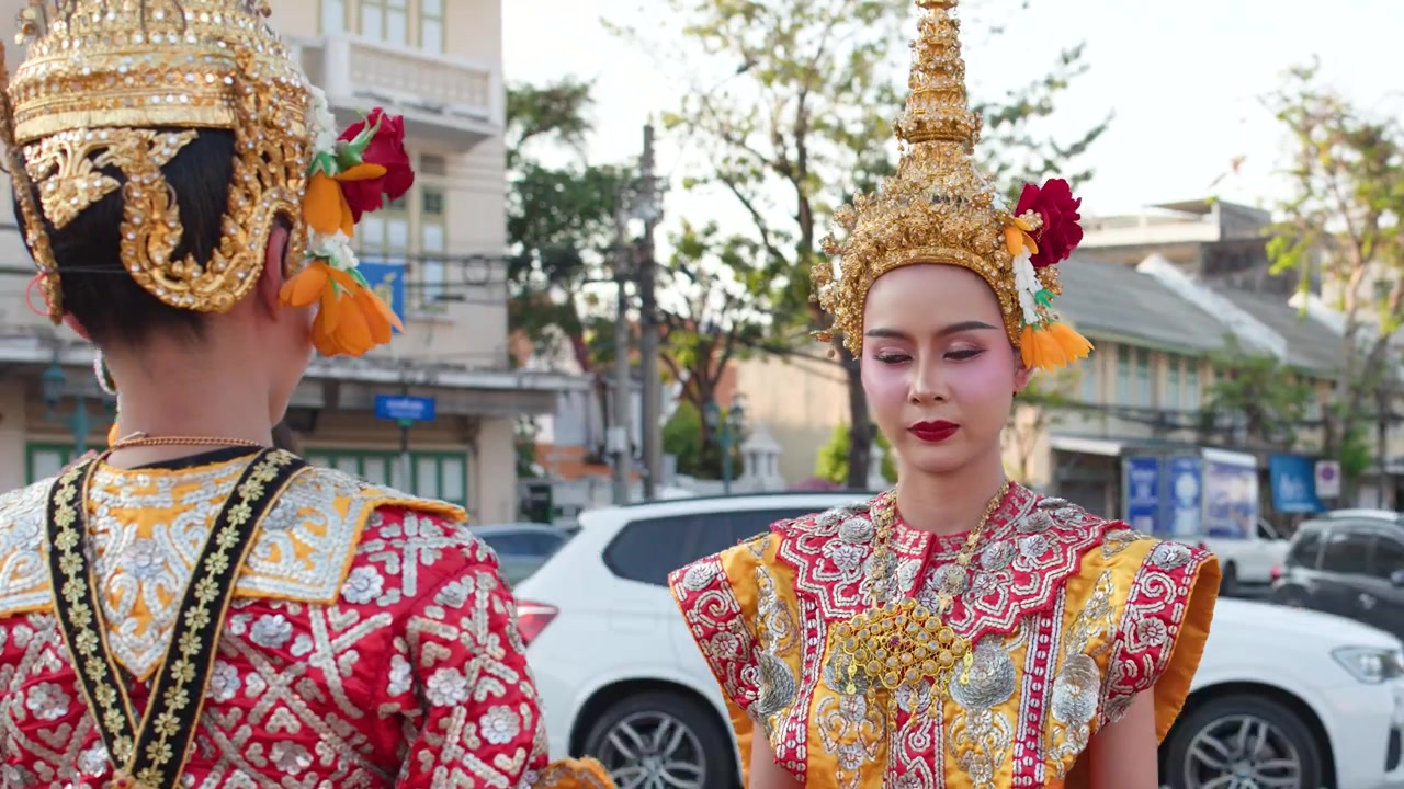 To thailandske Khon-dansere i udsmykkede traditionelle kostumer justerer hovedbeklædning udendørs i skarpt dagslys.