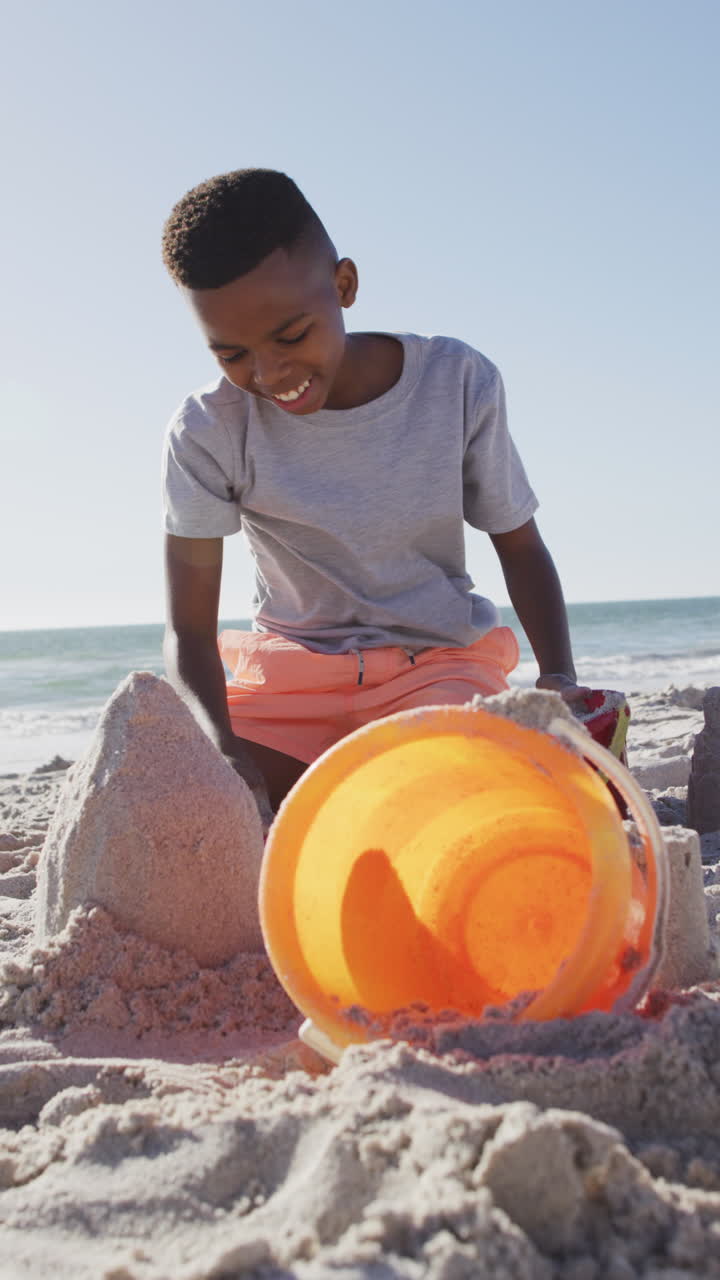 Vertical video of happy african american boy on beach playing with bucket