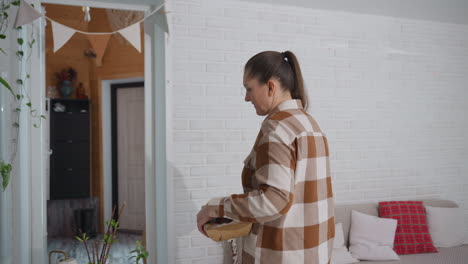 medium shot of woman gardener barefoot walking into cozy modern room carrying wooden basket then setting basket on round table near potted plants sofa sheer curtains and snowy view outside glass door