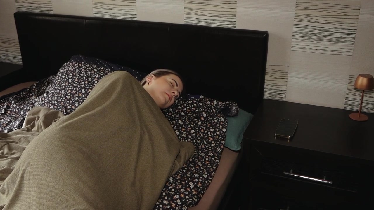 Donna sdraiata sotto una coperta a letto che si allunga verso uno smartphone su un comodino, creando una scena di vita tranquilla in camera da letto