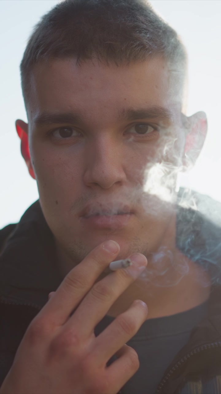 Primer plano de un hombre sosteniendo y fumando un cigarrillo con humo ascendiendo a su alrededor. El sujeto viste ropa casual, incluyendo una chaqueta, mientras el fondo está suavemente iluminado y desenfocado. La escena enfatiza.