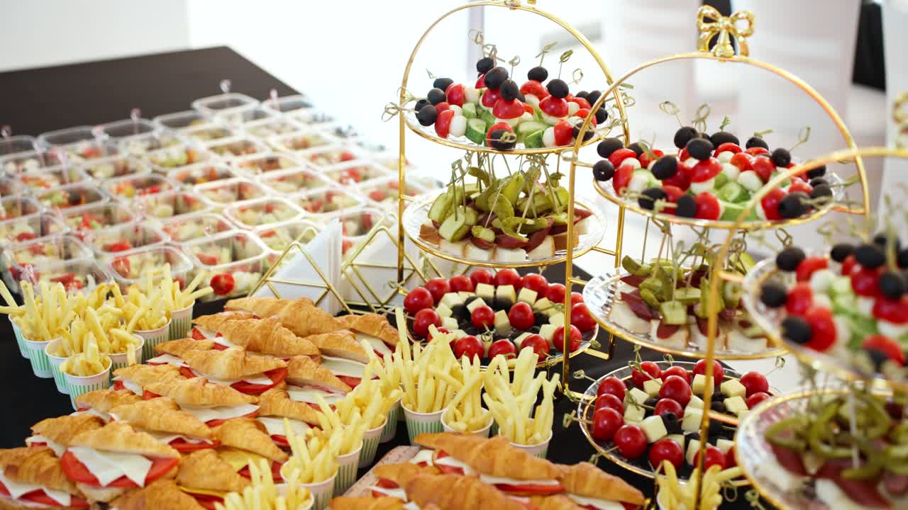 A slow dolly-in camera move pushes towards a delicious and varied catering buffet, showcasing croissant sandwiches, mini burgers, fries, and other appetizers for a special event.