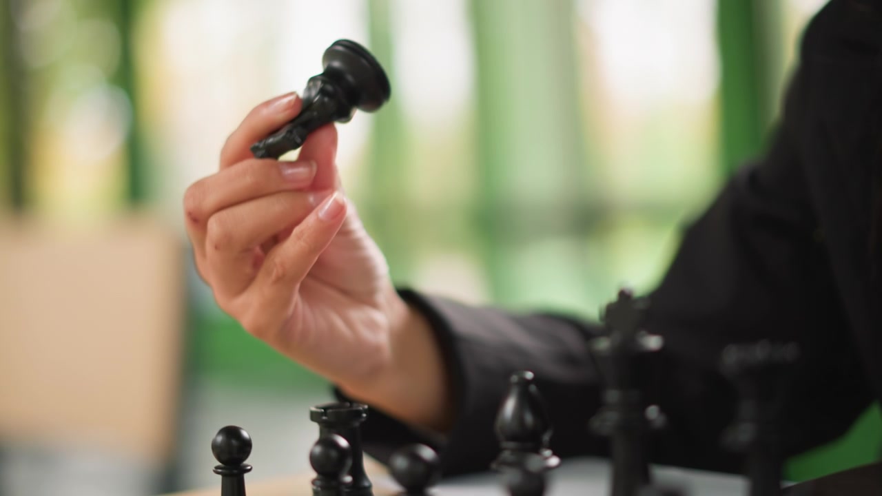 Primo piano della mano di una donna che muove pezzi degli scacchi neri sulla scacchiera. La messa a fuoco rimane nitida mentre la donna manipola vari pedoni e torri durante il gioco, con un'illuminazione soffusa che ne evidenzia i dettagli.