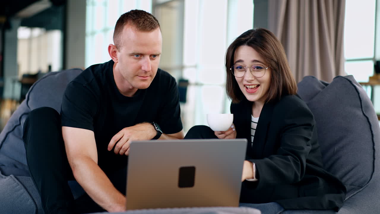 Man and woman sit in front of laptop looking at it focused. Colleagues laugh cheerfully watching video.