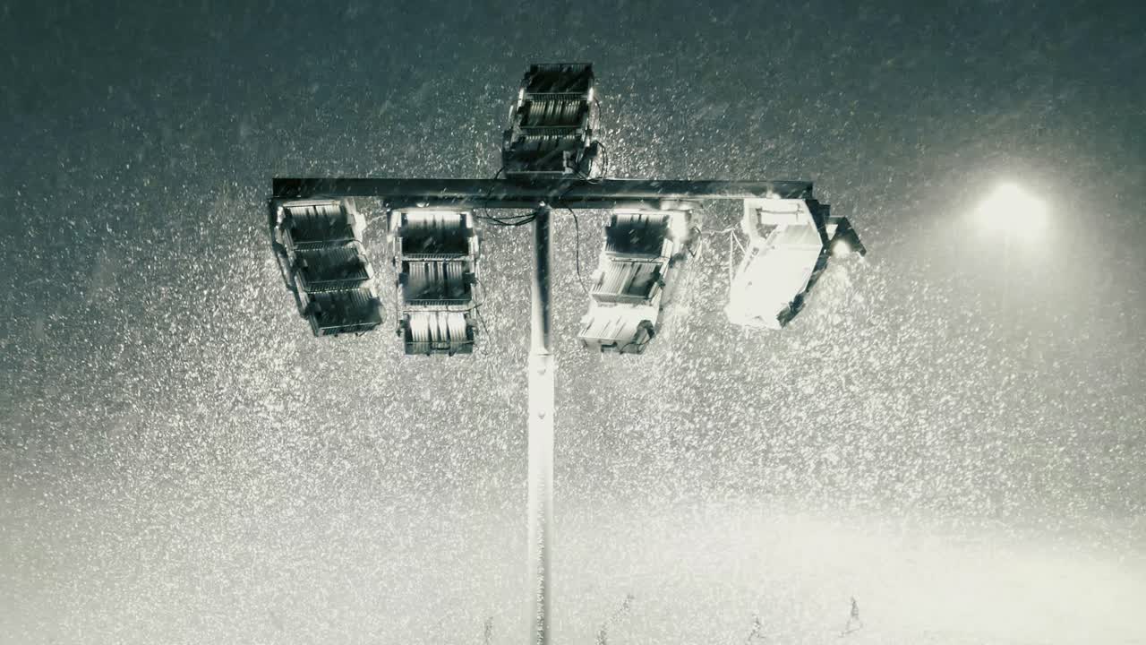 Circling aerial shot of stadium floodlight during snowfall blizzard. Stadium lighting illuminates falling snow while people are playing on football field. Spotlights illuminate the dark evening, night
