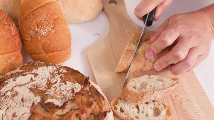 Close-up of hands using a knife to slice crusty baguette on a wooden cutting board.