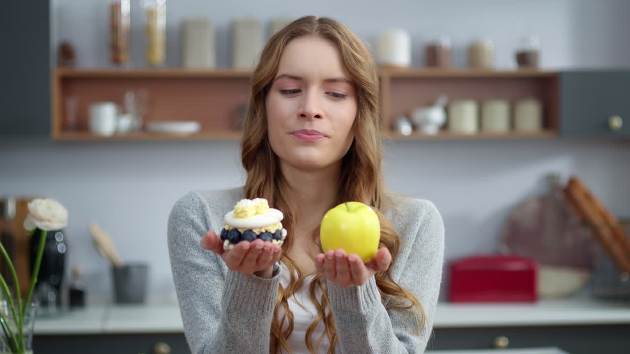 mujer sonriente mirando pastel y manzana en la cocina. chica que prefiere fruta fresca.