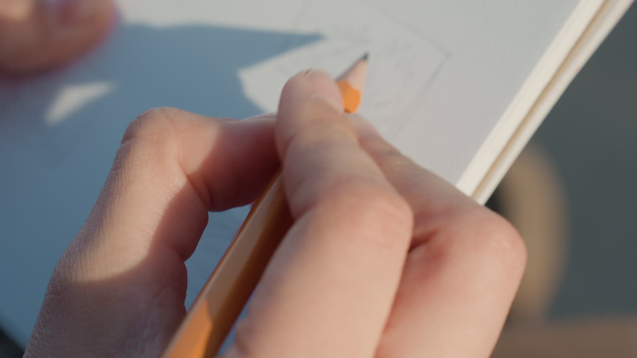 Primer plano de manos dibujando en papel, momento al aire libre soleado mostrando trazos de lápiz y sombra suave, artista refina pequeña composición con enfoque constante y sombreado cuidadoso, práctica íntima capturando