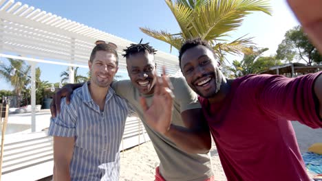 portrait of happy diverse male friends embracing on beach with beach house and palm trees