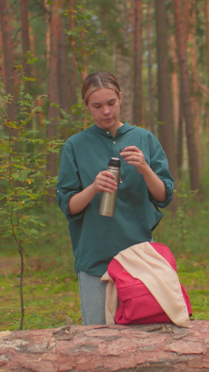 los excursionistas se detienen en un entorno sereno del bosque, uno llena la tapa del termo con agua, mientras que otros cepillan el cabello, ambos involucrados en rutinas personales, las mochilas descansan en un árbol caído