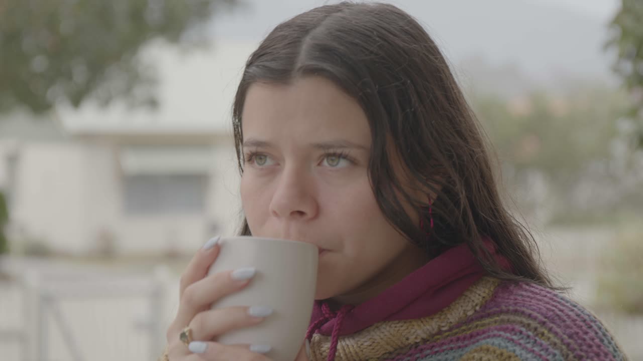Young Hispanic Latina woman sipping from a mug outdoors on chilly day, wrapped in colorful knit sweater, enjoying a quiet reflective moment