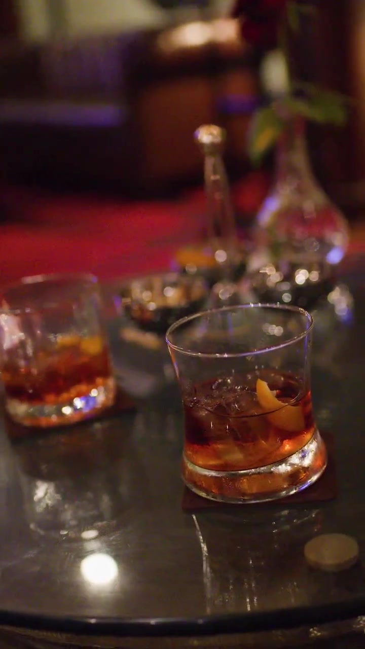 Bicchiere di whisky con ghiaccio su un tavolo da bar riflettente, illuminazione calda e soffusa, lenta carrellata in avanti della telecamera.