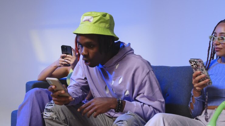 Studio Shot Of Young Gen Z Friends Sitting On Sofa Sharing Social Media Post On Mobile Phones 1