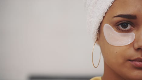 closeup portrait of beautiful woman after bath with towel on head puts patches under the eyes from wrinkles and dark circles. attractive female in bathrobe makes beauty treatments on the face at home.